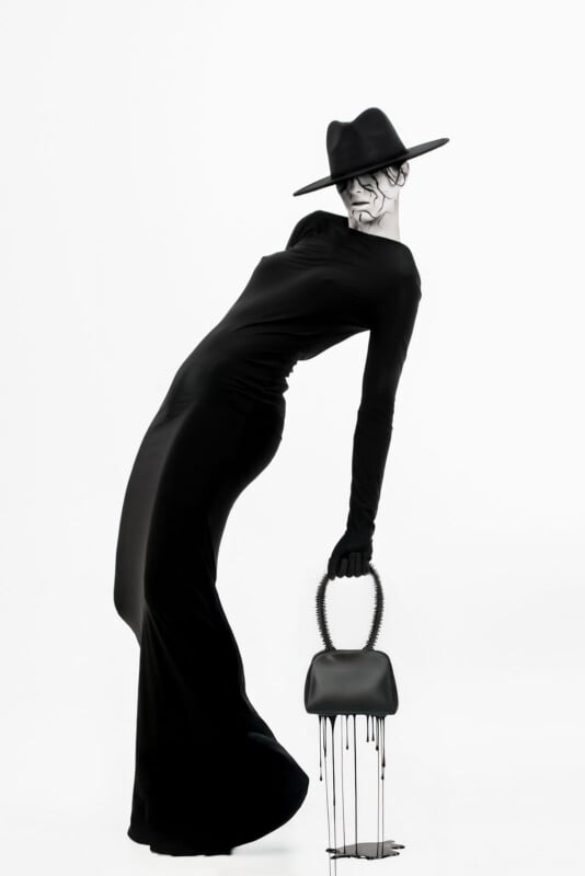 A person in a long, form-fitting black dress, wide-brimmed hat, and gloves leans dramatically backward, holding a handbag with dripping paint against a plain white background. Their face is partially shadowed and painted with dark lines.