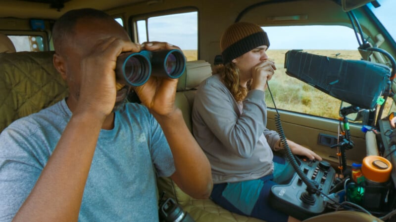 Two people sit in a vehicle, one looking through binoculars and the other speaking into a radio handset. They appear focused, possibly observing wildlife or conducting field research in a grassy outdoor area.