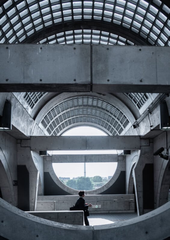 A person walks through a modern, concrete architectural space with large circular and arched windows, and a glass-paneled ceiling letting in natural light.