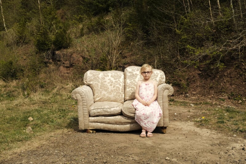 A young girl in a pink floral dress sits on an old, patterned sofa placed outdoors on dirt, with trees and bushes in the background. She is wearing glasses and looks directly at the camera.