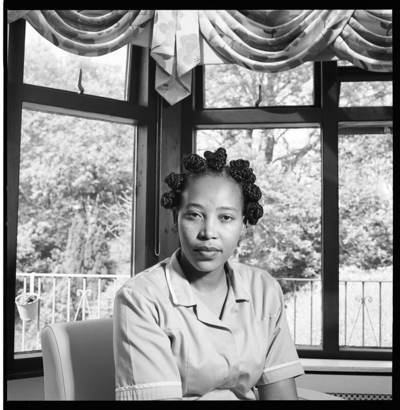 A woman wearing a uniform sits indoors in front of large windows with curtains, looking directly at the camera. Trees and a balcony are visible outside. The image is in black and white.