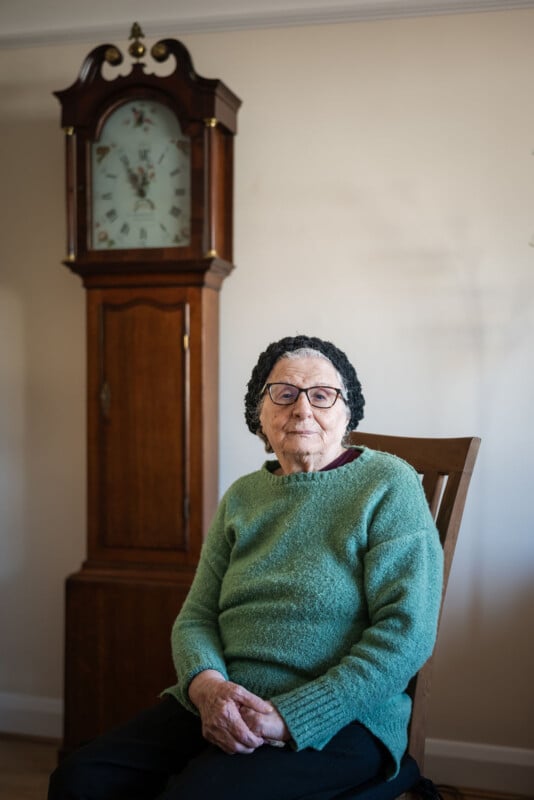 An elderly woman wearing glasses and a green sweater sits on a wooden chair in front of a tall, antique grandfather clock against a plain wall.