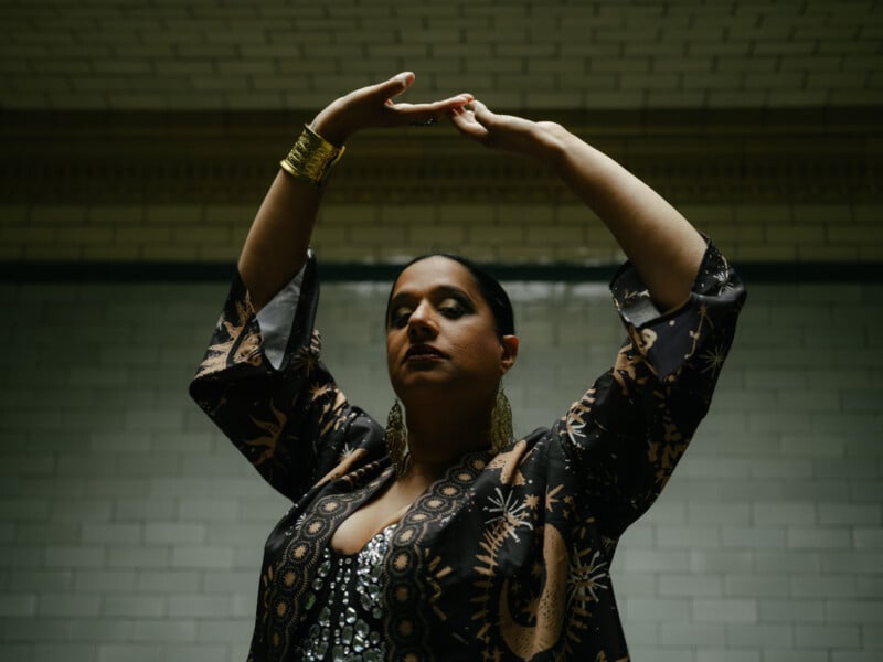 A woman wearing a patterned robe and large earrings raises her arms gracefully above her head, standing against a tiled wall in dim lighting.