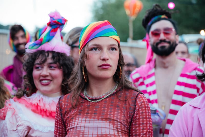 A group of people at a colorful outdoor event. The central person wears a rainbow headscarf and red patterned shirt, with others around them dressed in bright, playful costumes and makeup.