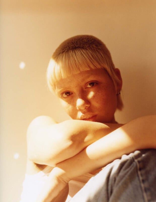 A person with short blonde hair and bangs sits with arms wrapped around their knees, gazing at the camera in warm, golden light. They wear a white top and light blue jeans. The background is softly blurred.