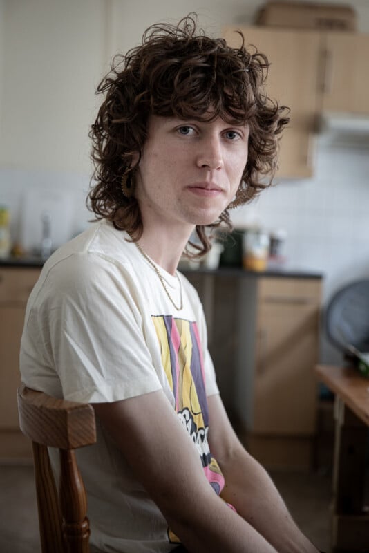 A person with curly brown hair and a light complexion sits on a wooden chair in a kitchen, wearing a graphic t-shirt and a gold chain, looking directly at the camera with a neutral expression.