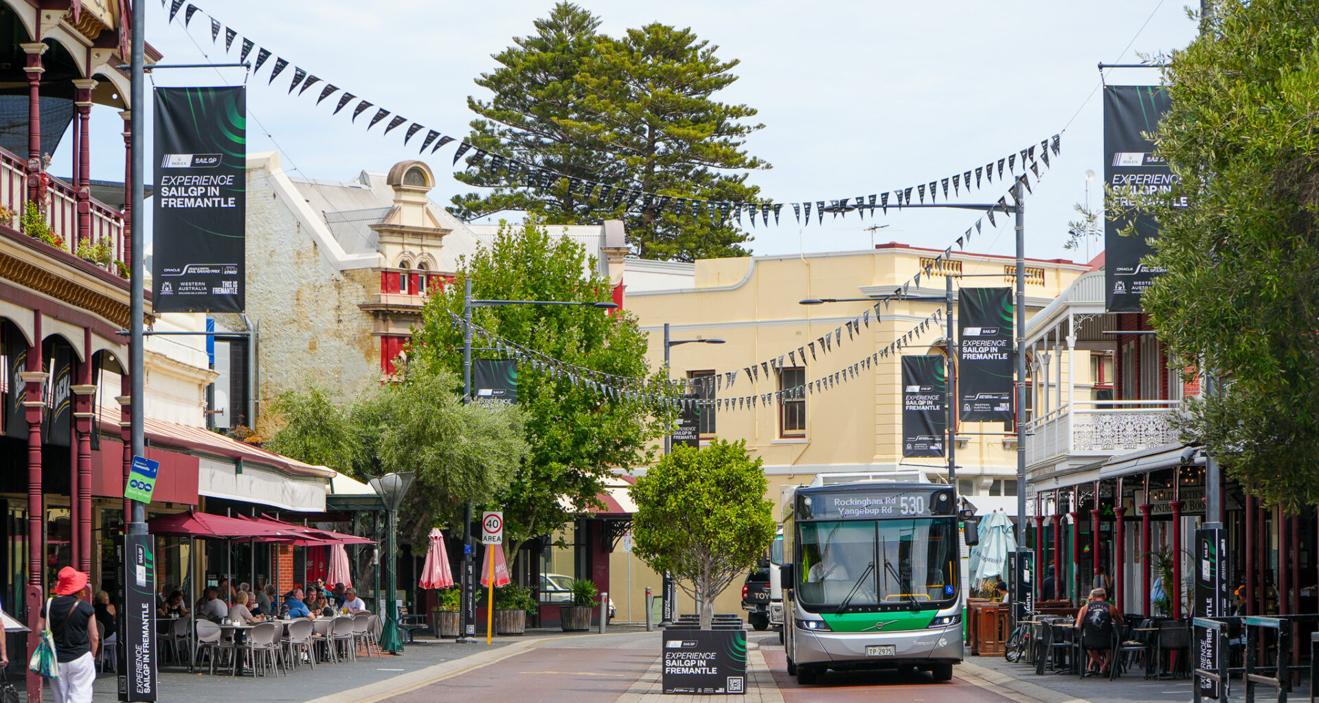 A green and white bus drives down a lively street lined with historic buildings, outdoor cafes, and hanging black flags. People are seated at tables, and banners promoting Fremantle hang above the street.