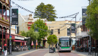 A green and white bus drives down a lively street lined with historic buildings, outdoor cafes, and hanging black flags. People are seated at tables, and banners promoting Fremantle hang above the street.