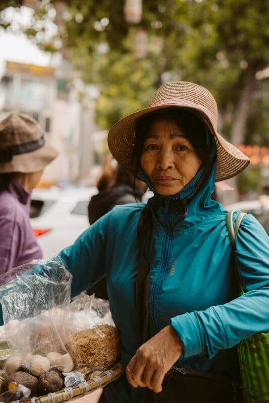 A woman wearing a wide-brimmed hat and blue jacket stands outdoors holding a basket with packaged food items. The background shows blurred people, trees, and cars on a city street.