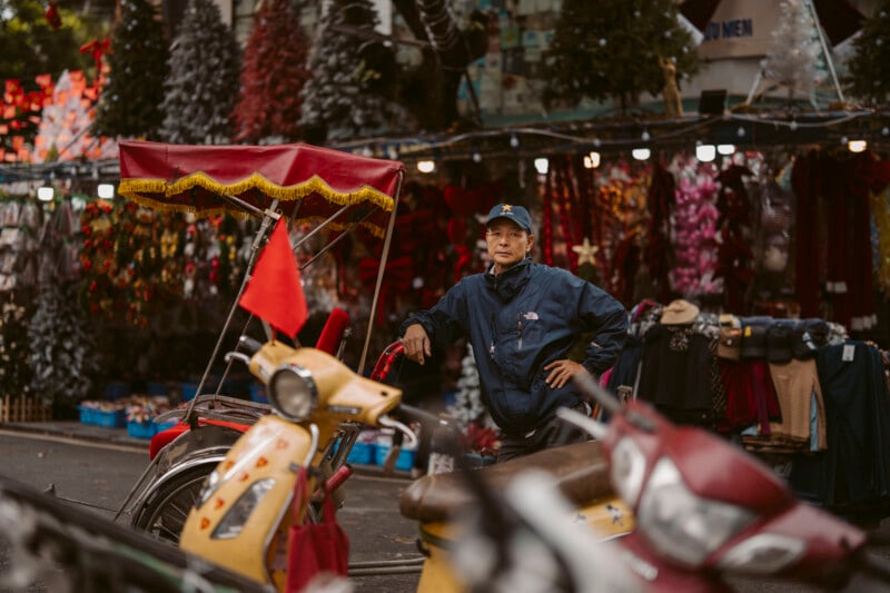 A person in a blue jacket and cap stands beside a decorated rickshaw on a busy street lined with colorful market stalls and artificial trees, with scooters parked in the foreground.