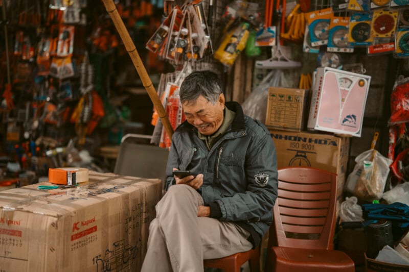 An elderly man sits on a plastic chair in a hardware shop, smiling while looking at his phone. Boxes and various tools hang around him, creating a busy and colorful background.
