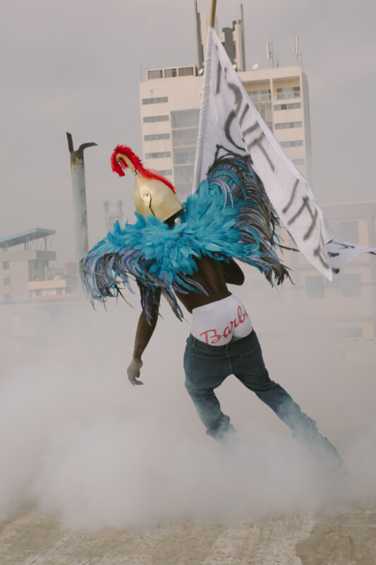 A person wearing a gold helmet, blue feathered wings, and "Barbie" underwear stands in jeans on a rooftop surrounded by smoke, holding a large white flag with buildings in the background.