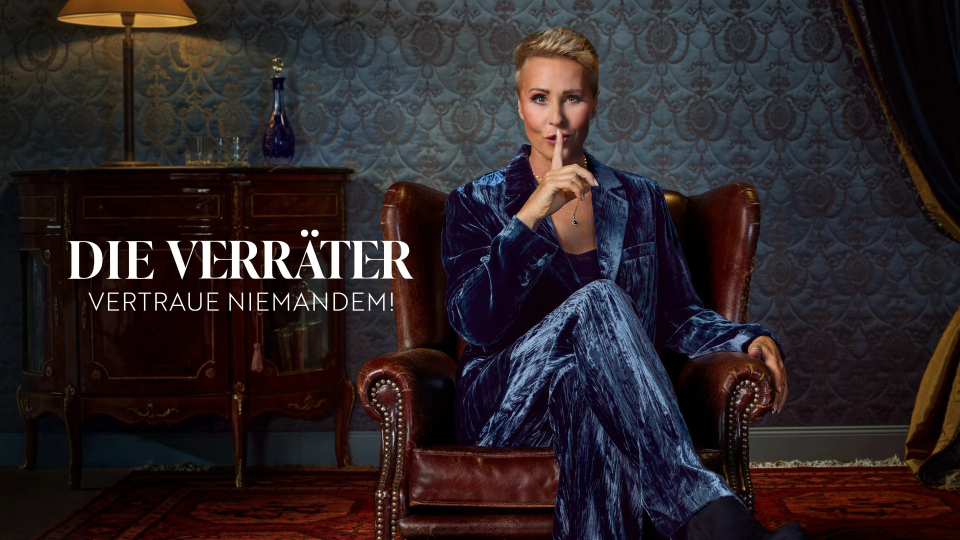 A person with short blonde hair sits in a leather armchair and holds a finger to their lips in a "shush" gesture, positioned in a dimly lit room next to the German text "Die Verr&auml;ter &ndash; Vertraue Niemandem!"