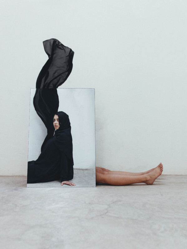 A mirror on the floor reflects a woman draped in flowing black fabric, with part of the fabric billowing upward; her bare legs extend from behind the mirror on a concrete surface against a plain wall.