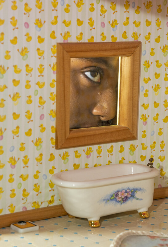A close-up of a person's face reflected in a small mirror above a mini bathtub with floral decoration, set against wallpaper with yellow duck illustrations.