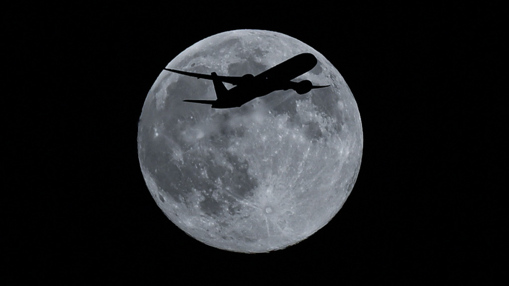 The moon is pictured against a black night sky with the dark silhouette of a passenger jet crossing its surface. Darker regions called lunar maria are also visible on its surface along with bright streaks of material cast outward from impact craters.