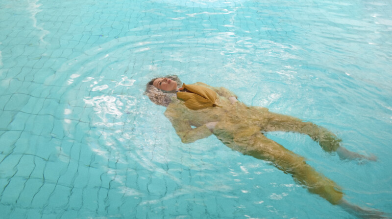 A person in a yellow outfit floats on their back in a swimming pool, with water rippling around them and their face partially above the water’s surface.