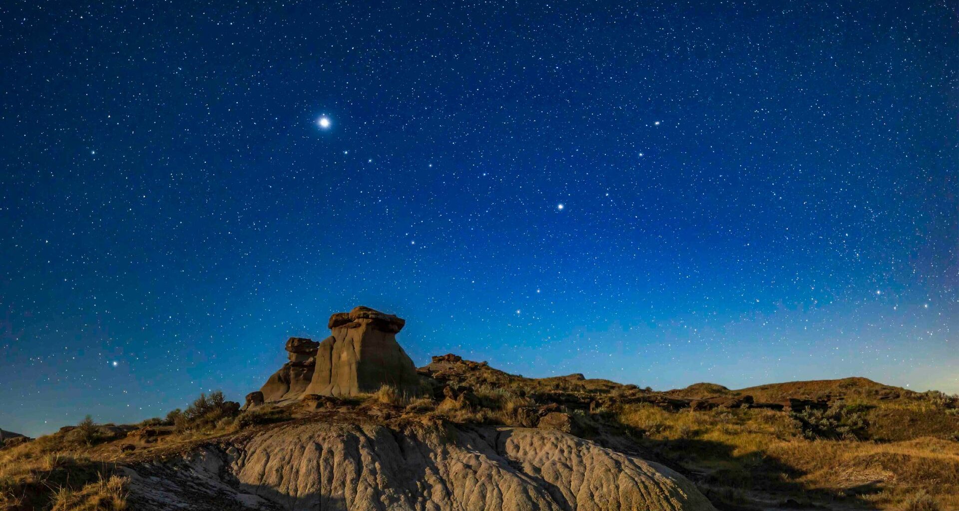 A glowing bright blue night sky full of white stars looks over a rocky landscape.