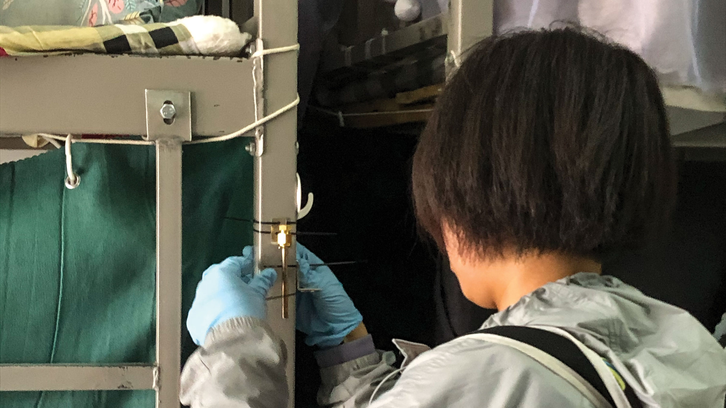 A person stands with their back toward the camera, attaching a small metal tube to a bed frame with zip ties.