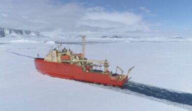 A large red icebreaker ship cuts through thick sea ice in a polar region, with snow-covered mountains and a cloudy sky in the background.