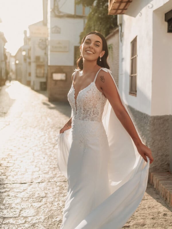 A smiling woman in a lace wedding dress stands on a sunlit cobblestone street, holding the skirt of her gown and looking joyfully at the camera.
