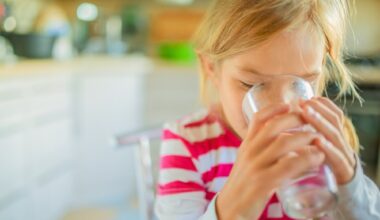 Beautiful smiling little girl drinking a glass of water against the background of the kitchen.