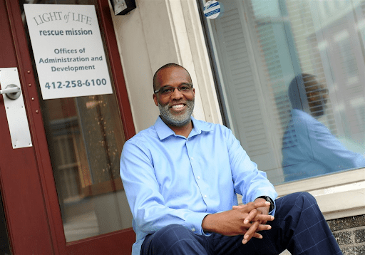 A man in a blue shirt sits and smiles in front of the entrance to the Light of Life Rescue Mission administration office.