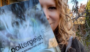 A person with curly hair holds up a book titled Poltergeist by Jocelyn Szczepaniak-Gillece outdoors near trees and a house.