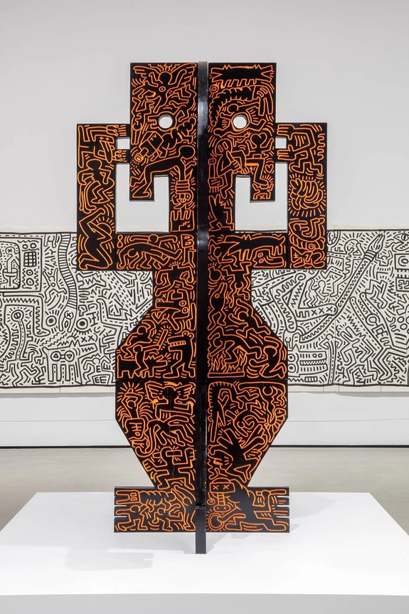 Contemporary sculpture with abstract, orange line patterns on black, displayed in an art gallery setting with mural in background