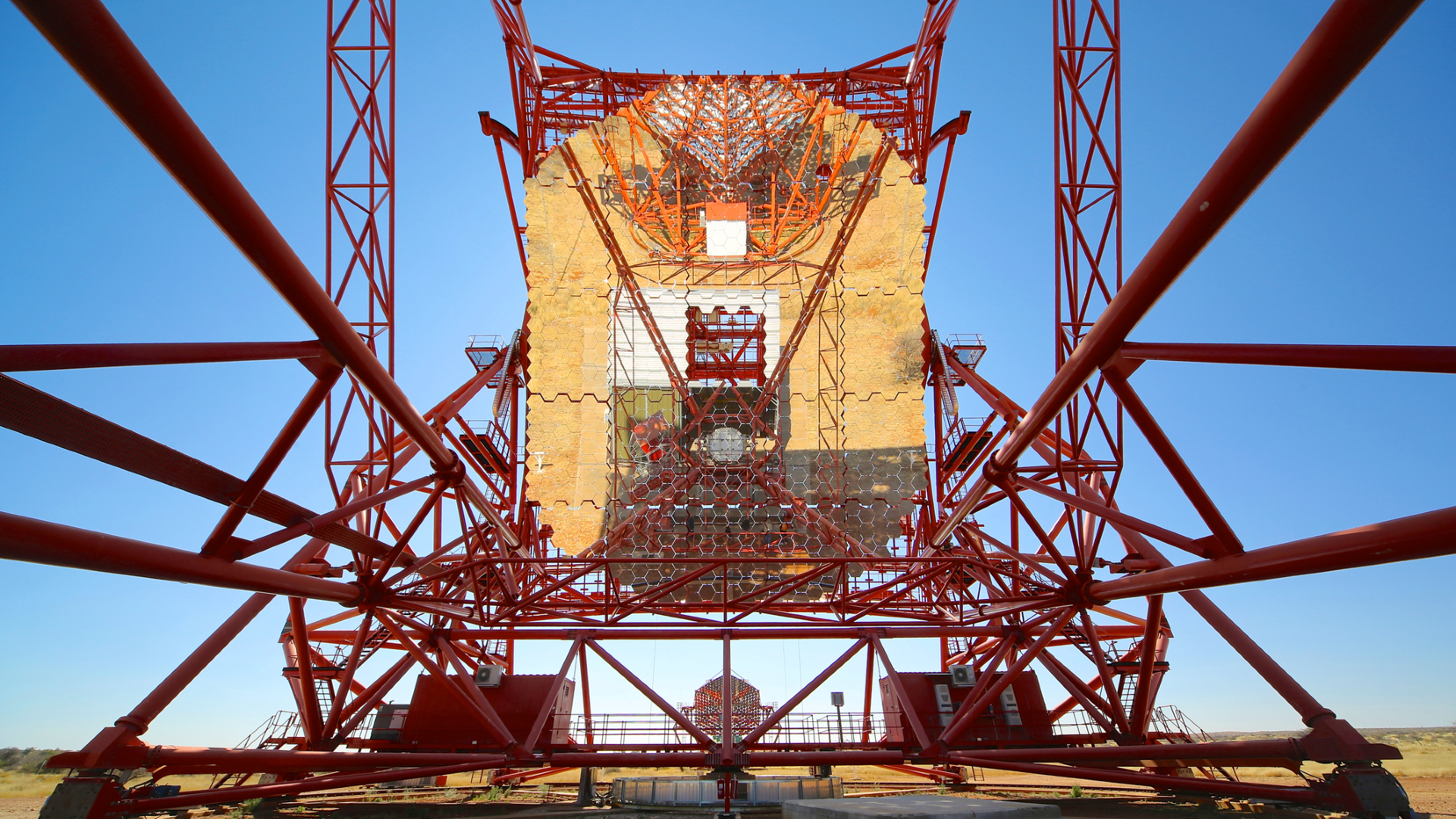 A golden telescope is held up by red scaffolding with a blue sky behind it.