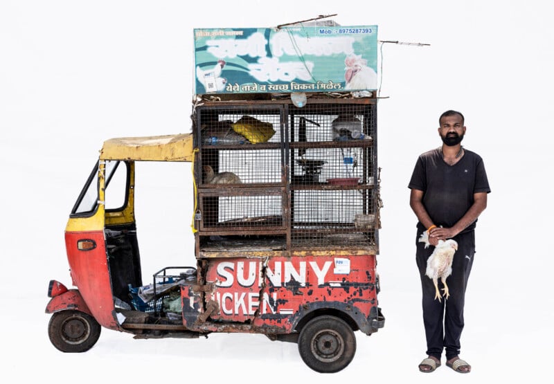 A man in black clothes stands holding a chicken beside a red and yellow auto rickshaw fitted with wire cages containing birds, against a plain white background. The vehicle reads "SUNNY CHICKEN" on the side.