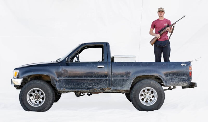 A man in a red shirt and hat stands in the bed of a blue pickup truck, holding a rifle. The truck is old and slightly dirty, and the background is plain white.