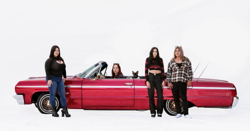 Four women and a small dog pose with a classic red convertible car against a plain white background. Two women stand on each side of the car, while one woman and the dog sit inside the car.