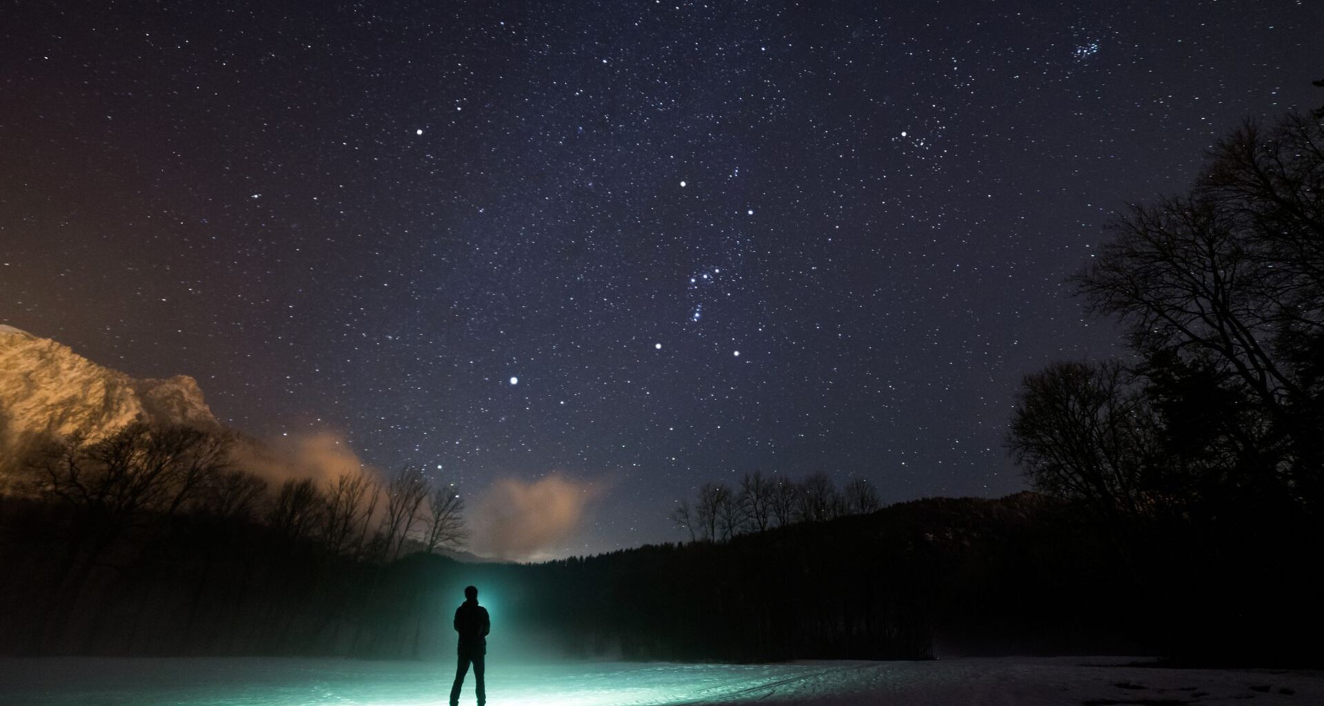 A silhouette of a person stands in front of a green flashlight beam on the edge of a lake with trees and hills in the distance all underneath a dark blue starry sky.