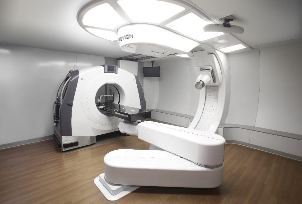A modern medical imaging and radiation therapy machine in a clean, well-lit room with wood flooring, designed to minimize human tissue damage from cosmic rays during treatment.