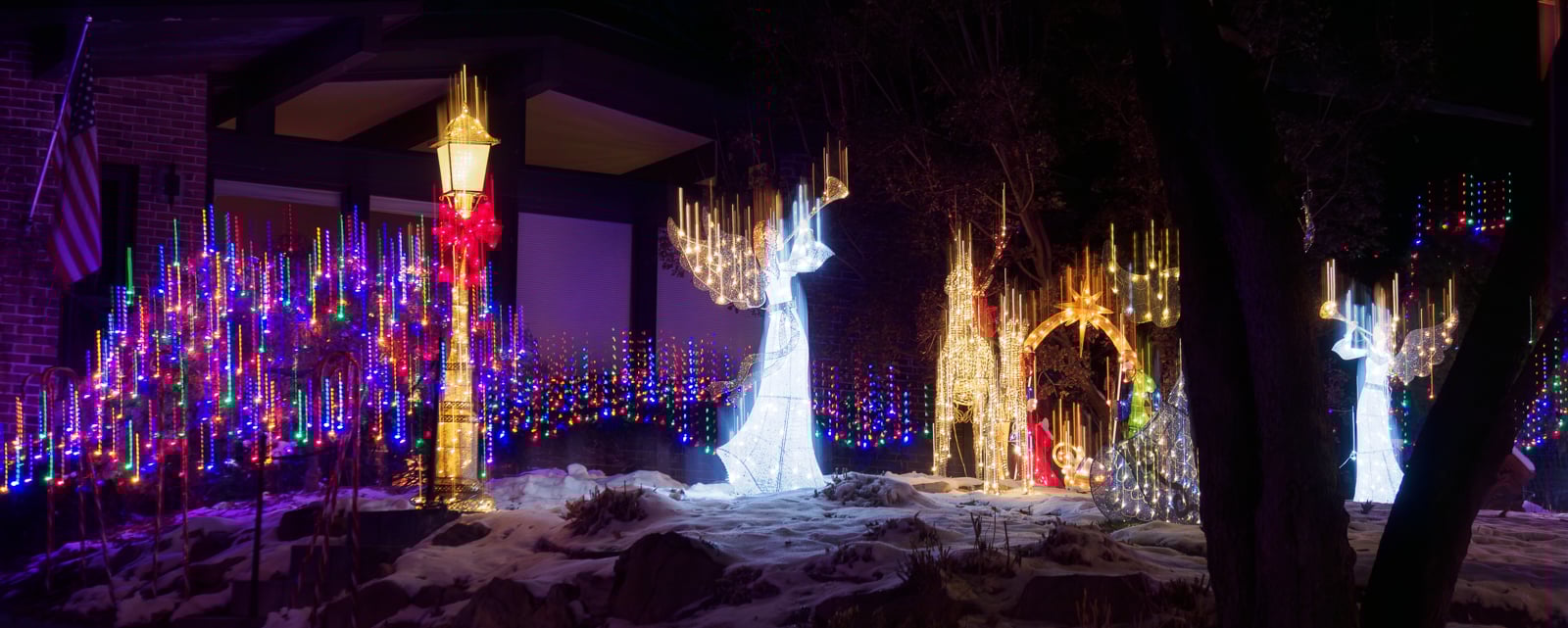 A snowy yard decorated with glowing angel and candelabra figures, a golden arch, and colorful string lights, creating a festive holiday scene at night in front of a house.