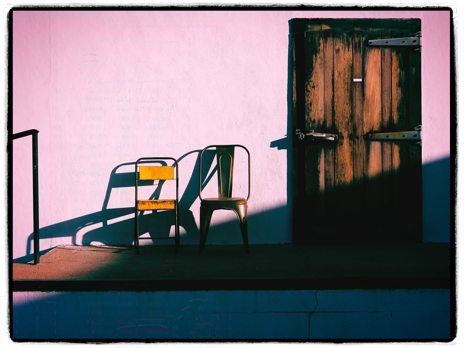 Two empty chairs, one yellow and one metal, cast long shadows against a pink wall next to a weathered wooden door partially in shadow. The scene is bathed in dramatic sunlight.