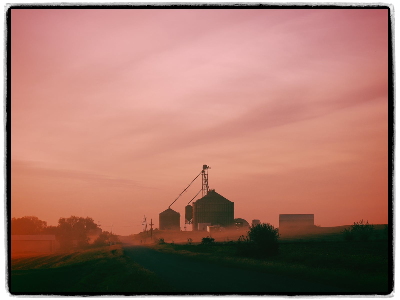 A rural landscape at sunset with soft pink sky, silhouettes of grain silos, a crane, and farm buildings, with a dusty road leading into the distance.