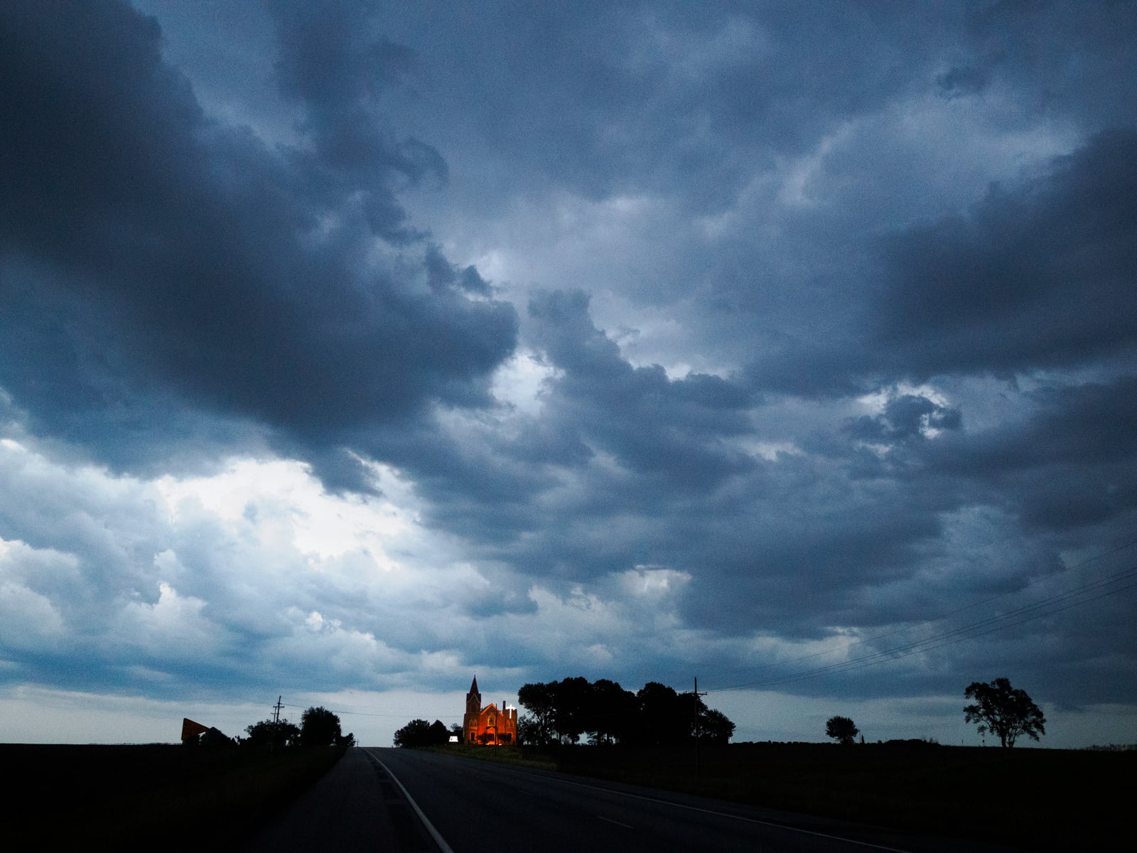 A dark, cloudy sky looms over a rural road at dusk. In the distance, a small, brightly lit building—possibly a church—stands surrounded by trees, creating a striking contrast against the dramatic sky.