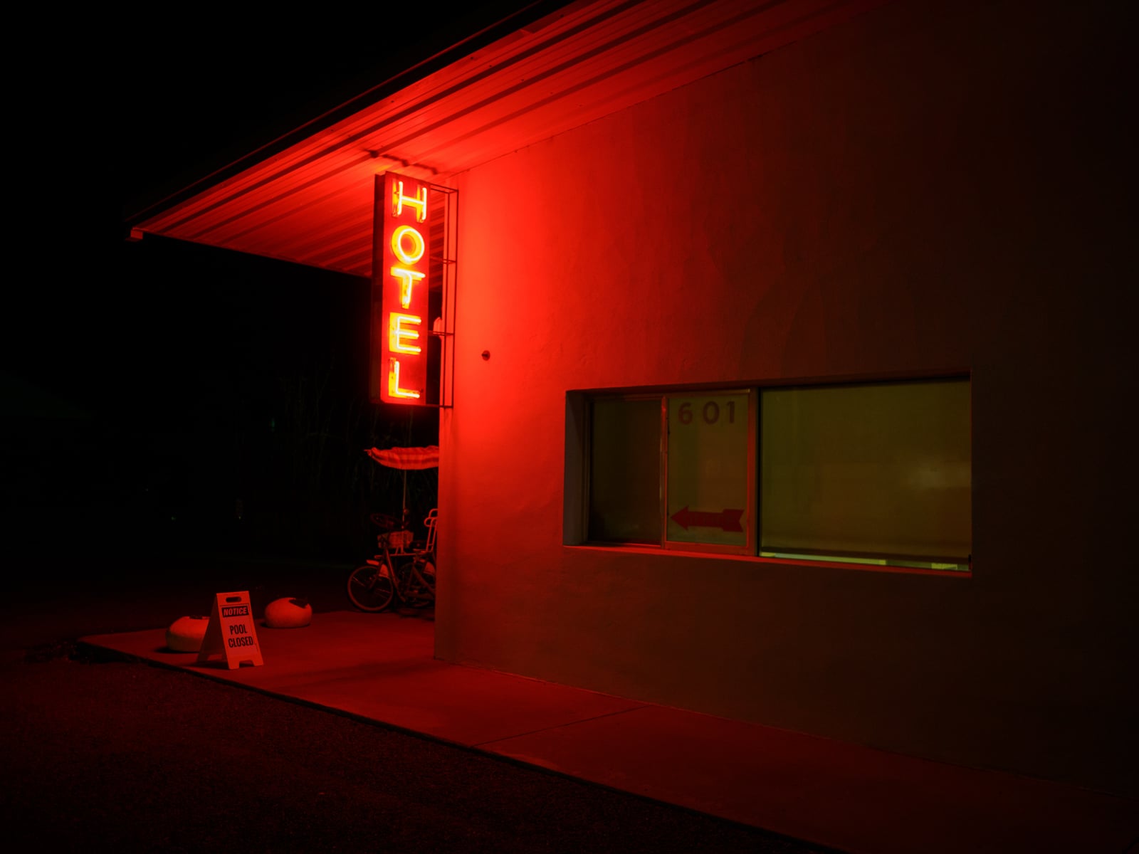 A bright red neon "HOTEL" sign glows at night above a building entrance. A sidewalk sign below reads "office open," and a lit window with the number 601 and an arrow is visible on the building.