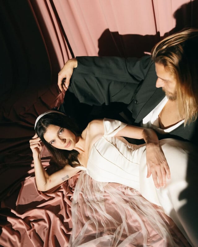 A woman in an off-shoulder white dress with tulle lies on pink fabric next to a man in a dark suit. They are bathed in soft sunlight, and she looks at the camera while he looks down, both appearing relaxed and elegant.