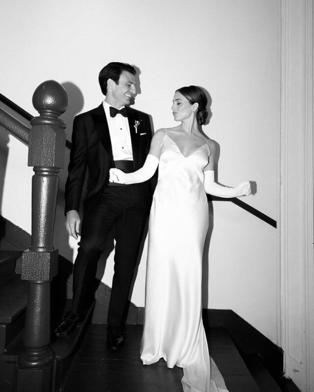 A couple dressed in formal attire stands on a staircase. The man wears a tuxedo and bow tie, while the woman wears a long satin gown and opera gloves. They smile and look at each other in a candid, elegant moment.