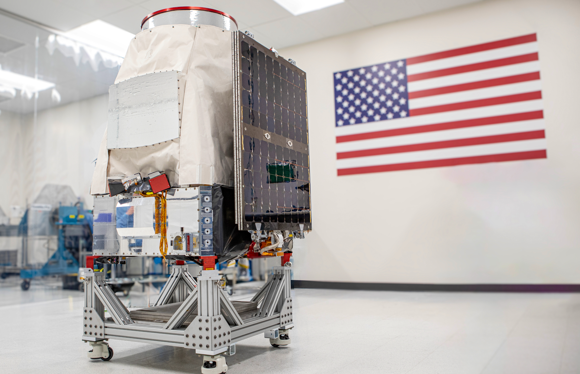 A covered metal probe sits on a cart in a clean room with the American flag hanging on the wall behind them