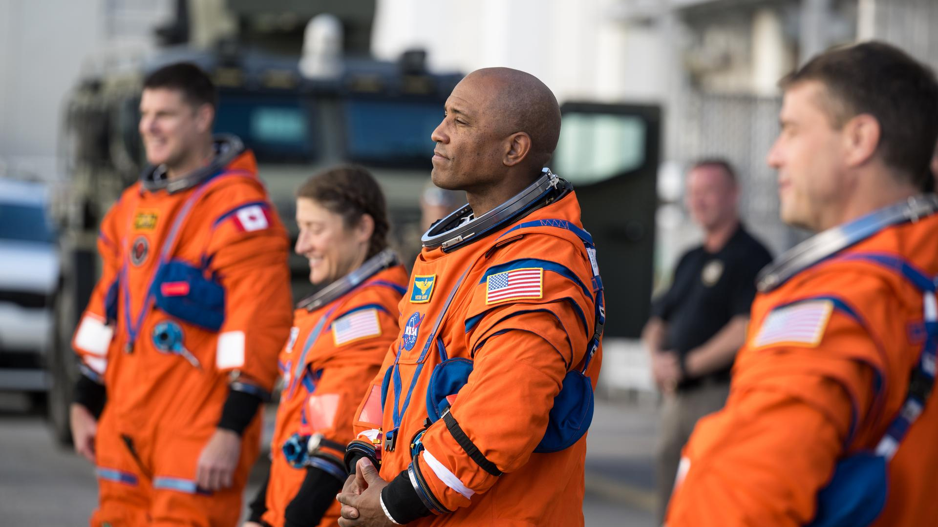 Four people in orange spacesuits address a crowd off camera to the left of the image