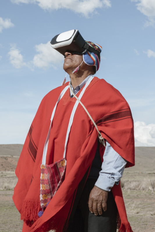 A person wearing traditional Andean clothing and a colorful hat uses a virtual reality headset outdoors, standing against a bright sky and natural landscape.