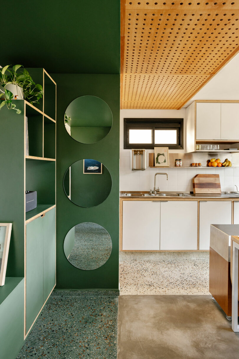 Modern kitchen with green accent wall featuring three circular mirrors, open shelving with plants, and white cabinets; wood details on ceiling and countertop.