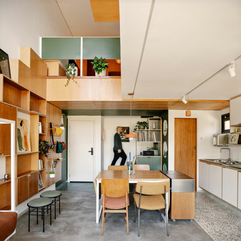 Modern open-plan apartment with wooden furniture, shelving, and a mezzanine level. A person walks near the white entrance door; kitchen and dining area are visible.