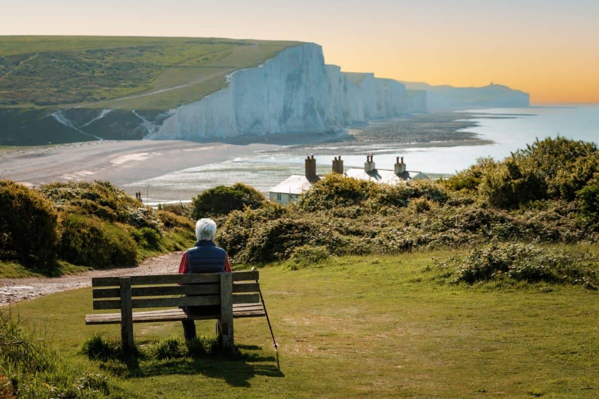 Rear view image depicting a senior man in his 70s sitting on a bench leading down to the iconic Seven Sisters cliffs on the coastline of East Sussex, UK. The man is wearing casual clothing - blue denim jeans, a red checked shirt, navy blue gilet. The man is having a rest from hiking and his hiking pole is leaning up against the bench.