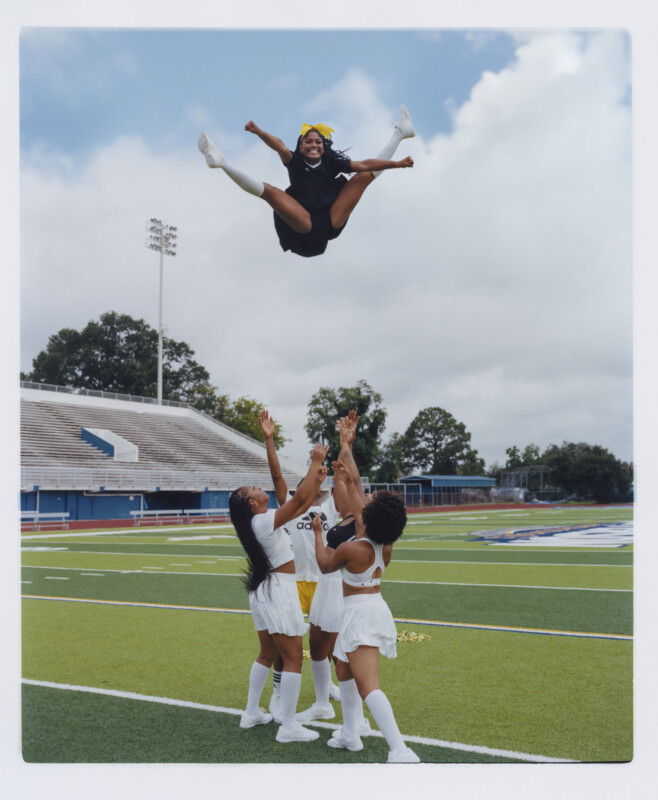 Four cheerleaders in white uniforms lift another cheerleader in black high into the air on a football field, with empty bleachers and a cloudy sky in the background. The airborne cheerleader wears a yellow bow and smiles.