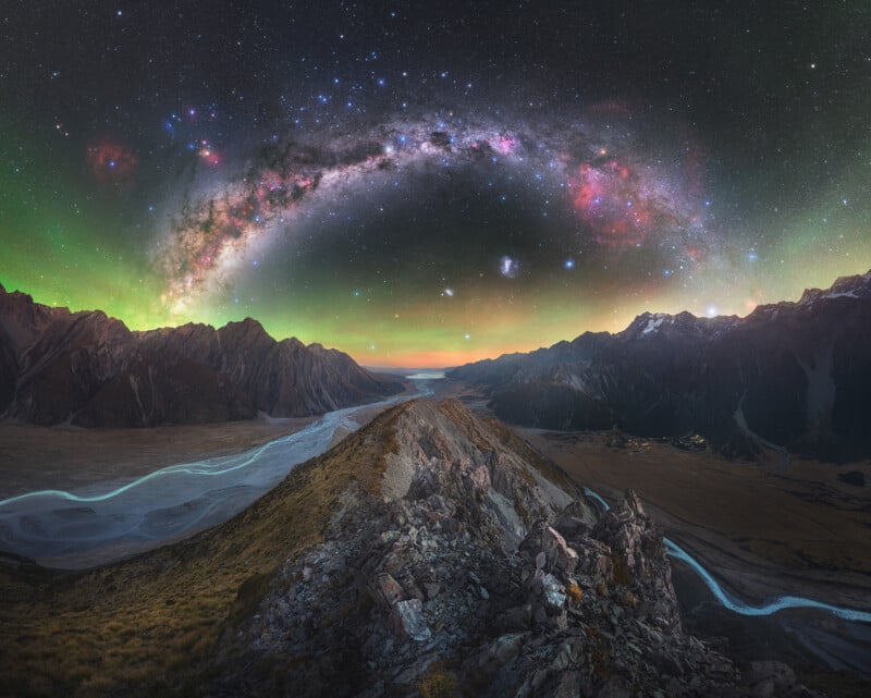 A rocky mountain ridge leads to a winding river valley at dusk, with a vivid, colorful aurora and the Milky Way arched across a star-filled night sky above. Snow-capped peaks line the horizon.